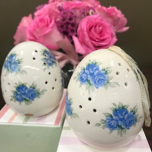 Op de afbeelding: Twee witte, eivormige keramische ornamenten met blauwe rozenontwerpen en kleine gaatjes. De ornamenten staan op een roze-wit gestreepte doos.