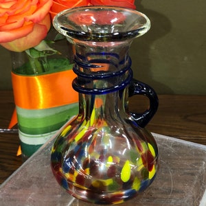 Könnte beinhalten: Eine kleine, klare Glasvase mit einem blauen Wirbelmuster und einem Henkel. Die Vase ist mit buntem, gesprenkeltem Glas in Rot-, Gelb- und Blautönen verziert.
