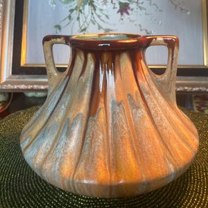 Puede incluir: Un jarrón de cerámica con base ancha y dos asas. El jarrón tiene una superficie texturizada con una mezcla de colores naranja, marrón y gris azulado. El borde interior es de color marrón oscuro. El jarrón es probablemente para fines decorativos.