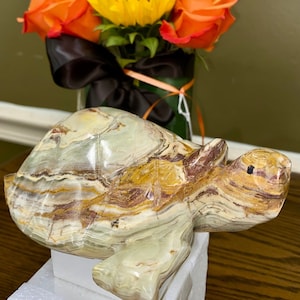 May include: A carved stone turtle figurine with a multi-colored pattern of green, brown, and yellow. The turtle is resting on a white surface, with a vase of orange roses and a yellow sunflower in the background.