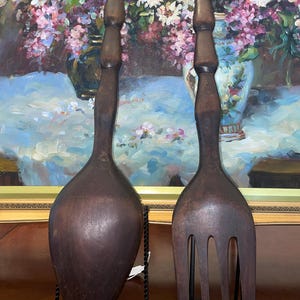 Könnte beinhalten: Zwei dunkelbraune Servierutensilien aus Holz. Eines ist ein großer Löffel, das andere eine Gabel. Beide haben lange, gedrechselte Griffe und sind wahrscheinlich Vintage oder antik. Sie werden vor dem Hintergrund eines Blumenbildes präsentiert.