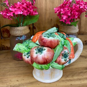 May include: A white ceramic teapot shaped like a basket overflowing with colorful vegetables. The teapot features a red onion, green peppers, carrots, and tomatoes. The teapot has a handle and a spout.