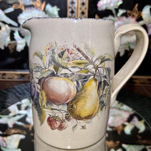 May include: Cream-colored ceramic pitcher with a fruit illustration. The pitcher features a handle and a spout. The fruit illustration includes a peach, pear, strawberries, and plums. The pitcher has a dark blue trim around the rim.
