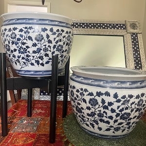 May include: Two white and blue ceramic planters with floral designs. One planter sits on a black wooden stand. The planters have a wide rim with a blue wave pattern. The floral design is a dark blue color.