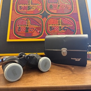 May include: Black binoculars with white lens caps and a brown leather strap sit on a wooden surface. A black Tasco case is next to the binoculars. Behind them is a framed artwork with red, yellow, and black geometric designs.