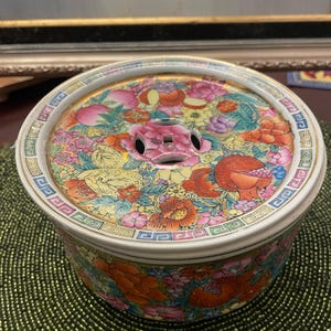 Pode incluir: Um recipiente redondo de cerâmica com tampa e um design floral vibrante. A tampa apresenta uma peça central floral rosa com três orifícios, rodeada por flores e frutas coloridas. O recipiente tem uma borda branca com uma borda geométrica.