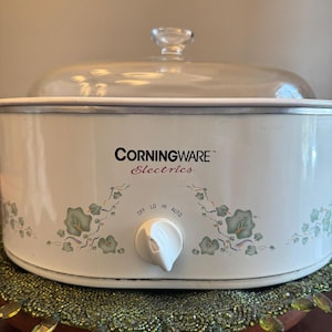 May include: A vintage CorningWare Electrics slow cooker with a clear glass lid. The white appliance features a control knob with settings for off, low, high, and auto. It is decorated with green floral patterns.