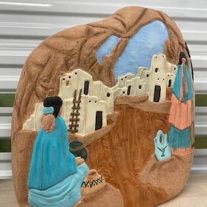 Op de afbeelding: Een keramische sculptuur van een scène uit een indianendorp. De sculptuur is geschilderd in tinten bruin, blauw en wit, en toont een groep mensen in traditionele kleding. De sculptuur is gevormd als een rots en heeft een getextureerd oppervlak.