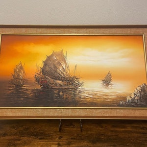 May include: A framed oil painting depicting three traditional Chinese junks sailing on a calm sea at sunset. The sky is a vibrant orange and yellow, and the water is a deep blue. The junks are detailed with sails and rigging, and the painting is framed in a simple brown frame.