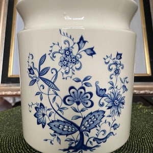 Puede incluir: Un recipiente blanco de cerámica para utensilios de cocina con un diseño floral azul. El recipiente cilíndrico presenta una parte superior acampanada y está decorado con flores, hojas y enredaderas azules detalladas. El diseño recuerda a la porcelana clásica azul y blanca.