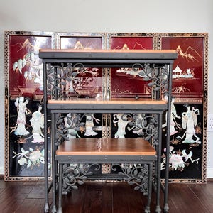 May include: A set of three black wrought iron nesting tables with wood tops. The tables feature a floral design and are in front of a black and red screen.