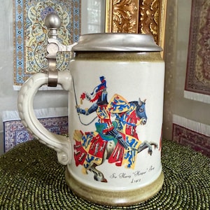 May include: A ceramic beer stein with a metallic hinged lid and a curved handle. The body features a colorful illustration of a knight on horseback in armor, with the text Sir Harry Hotspur Percy d. 1403 printed below the graphic design.