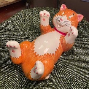Op de afbeelding: Een keramieken kattenbeeldje in een speelse pose, liggend op zijn rug met de poten omhoog. De kat heeft oranje vacht met een witte buik en poten, roze oren en een roze halsband. Het gezicht van de kat heeft een glimlachende uitdrukking.