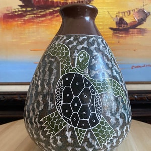 Puede incluir: Un jarrón decorativo hecho a mano con una parte superior marrón oscuro y un cuerpo verde y negro. El jarrón presenta un diseño detallado de tortuga con detalles en blanco. El jarrón probablemente está hecho de cerámica o un material similar.