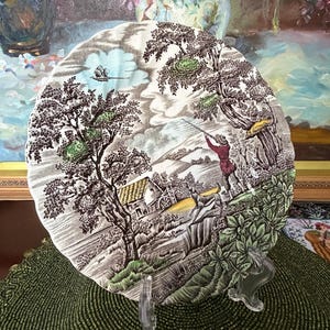 May include: A decorative plate with a pastoral scene in shades of brown, green, and white. The plate features a detailed illustration of a man, trees, a house, and a boat. The plate is displayed on a clear stand.