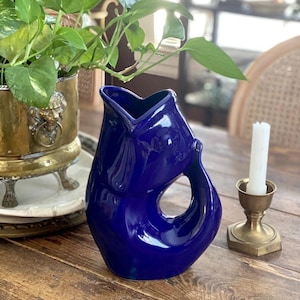 May include: A glossy, deep cobalt blue ceramic pitcher shaped like a fish. The pitcher features a wide, open mouth at the top that serves as the pouring spout, with a curved handle forming the tail section of the fish body design.
