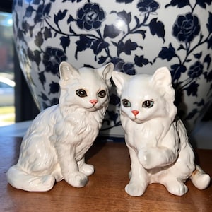 Może przedstawiać: Dwie białe ceramiczne figurki kotów ze szczegółowym futrem i wyrazistymi oczami. Jeden kot siedzi wyprostowany, a drugi podnosi łapę. Tło przedstawia duży wazon z niebiesko-białym wzorem kwiatowym.