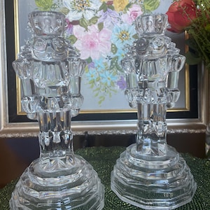 May include: Two clear glass nutcracker candle holders. Each candle holder has a detailed design, including a crown and uniform. The bases are multi-faceted. The background includes a painting of flowers.