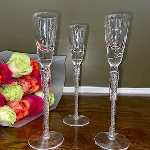May include: Three clear crystal champagne flutes with intricate stem detailing. The flutes are arranged on a wooden surface, with a bouquet of roses in the background. The stems have a twisted design.