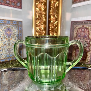 May include: A light green glass sugar bowl featuring a vertical ribbed pattern and two side handles. The bowl has a scalloped rim design and a clear, translucent finish, showcasing the pressed glass construction with a smooth, rounded base.
