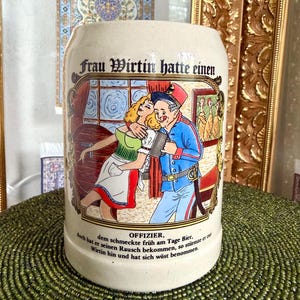 May include: A beige ceramic beer stein featuring a colorful illustration of a woman and an officer in uniform. The text reads: Frau Wirtin hatte einen OFFIZIER, dem schmeckte früh am Tage Bier, doch hat er seinen Rausch bekommen, so stürmte er zur Wirtin hin und hat sich wüst benommen.