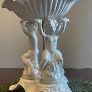 May include: A white ceramic centerpiece bowl with a scalloped rim, supported by two cherubic figures. The figures are holding the bowl aloft, standing on a decorative base. The bowl and figures have a glossy finish.
