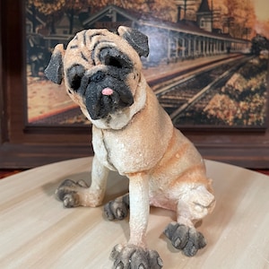 Könnte beinhalten: Eine Figur eines braunen und schwarzen Mops, die auf einer hellen Holzoberfläche sitzt. Die Figur hat ein detailliertes Gesicht und Pfoten.