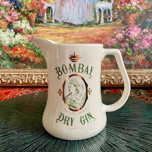 Puede incluir: Jarra de cerámica blanca con un logotipo de Bombay Dry Gin en oro y verde. El logotipo presenta una corona, un retrato de una mujer y el texto "Distilled London Dry Gin Since 1761".
