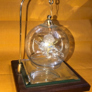 Może przedstawiać: Szklana ozdoba z pozłacanym aniołem w środku, umieszczona na drewnianej podstawce z lustrzaną podstawą.