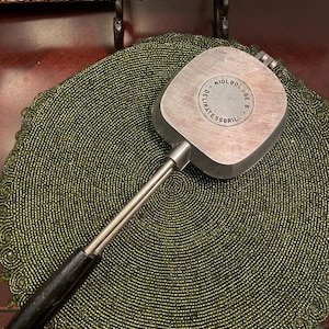 Puede incluir: Una plancha de gofres de metal vintage con una superficie de cocción cuadrada y un mango largo. La plancha tiene un mango de madera negro y el texto "NIOLBOLAGE S. DELIKATESSRILL" es visible en la superficie de cocción.