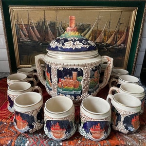 Puede incluir: Un tazón grande de ponche de cerámica blanca con un diseño azul y dorado. El tazón está decorado con imágenes de castillos y vides. Está rodeado de 10 tazas de cerámica más pequeñas con diseños similares. Las tazas son todas blancas con acentos azules y dorados.