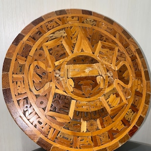May include: A round wooden wall hanging featuring an intricate design of a sun with a face in the centre. The design is crafted from various pieces of wood, creating a mosaic effect.