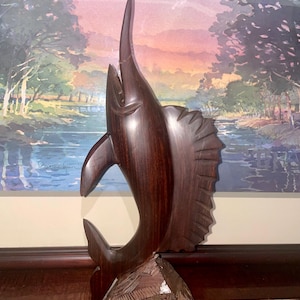 May include: A carved wooden sculpture of a marlin fish, showcasing a dark brown finish. The fish is depicted leaping upwards, with detailed fins and a textured base. The sculpture is set against a backdrop of a landscape painting.