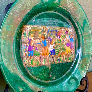 Könnte beinhalten: Ein grüner Keramikteller mit erhöhtem Rand und einem transparenten Glaseinsatz. Der Einsatz enthält eine farbenfrohe handgemalte Illustration von Menschen und Tieren.