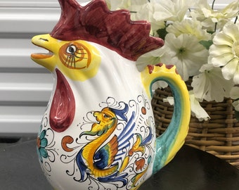 Puede incluir: Una jarra de cerámica blanca con un diseño de gallo. La jarra presenta una cresta roja, un pico amarillo y un diseño pintado colorido de un pájaro y elementos florales. El asa está pintada con rayas amarillas, azules y verdes.