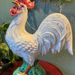 Puede incluir: Una estatua de gallo de cerámica blanca con cresta y barbilla rojas y pico amarillo. El gallo se alza sobre una base verde y azul. La estatua está sobre una mesa roja.