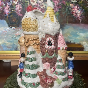 Può includere: Un villaggio natalizio in ceramica con un design a forma di casa di pan di zenzero. La casa presenta glassa bianca, decorazioni di caramelle e un orsacchiotto. Due soldatini si trovano su entrambi i lati della casa. Il villaggio è su un tappetino verde.