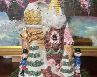 May include: A ceramic Christmas village with a gingerbread house design. The house features white icing, candy accents, and a teddy bear. Two toy soldiers stand on either side of the house. The village sits on a green mat.