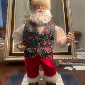 Peut inclure: Figurine du Père Noël avec barbe blanche et bonnet garni de fourrure. Il porte un gilet à motifs sur une chemise blanche, un pantalon en velours rouge et des bottes noires. Décoration de Noël festive.