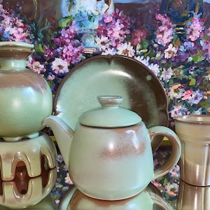 Puede incluir: Una tetera de cerámica verde y marrón glaseada con una tapa a juego, una taza de té a juego y un platillo a juego. La tetera está sentada sobre un soporte a juego con tres patas. El conjunto está frente a una pintura de flores.
