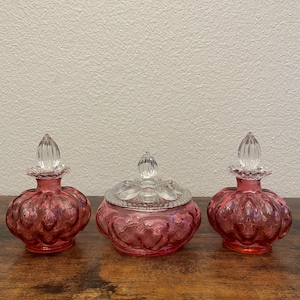 Puede incluir: Tres artículos de tocador antiguos de cristal de color rubí. Dos frascos de perfume con tapones transparentes en forma de lágrima flanquean un pequeño tarro con tapa. El cristal tiene un diseño texturizado y acanalado. Los artículos están dispuestos sobre una superficie de madera oscura.