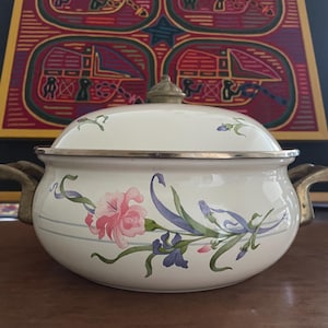 May include: Cream-colored enamelware pot with lid, featuring a floral design in pink, purple, and green. The pot has brass-toned handles and rim. A decorative piece for the kitchen.