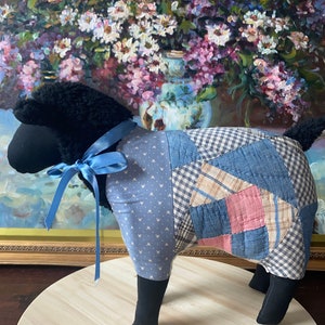 Puede incluir: Una figura de oveja hecha a mano con cabeza y patas negras, y un cuerpo estilo patchwork en tonos azules, rosas y beige. Una cinta azul está atada alrededor del cuello. La oveja se exhibe sobre una superficie de madera.