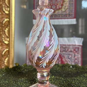 Peut inclure: Flacon de parfum en verre rose avec bouchon assorti. Le flacon a un corps en forme de poire avec des motifs blancs en forme de plumes. Le flacon repose sur une base ronde. L'arrière-plan comprend un tapis décoratif et un miroir doré.