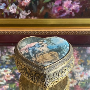 May include: A heart-shaped music box with a gold finish and intricate floral designs. The lid features a painted scene of a couple in a garden setting.
