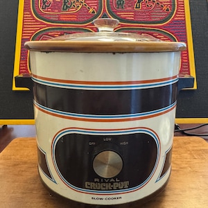 Peut inclure: Mijoteuse Rival Crock-Pot vintage. L'appareil a un extérieur blanc et marron avec des rayures bleues et rouges. Le panneau de commande est noir avec un bouton argenté et les mots "RIVAL CROCK-POT SLOW-COOKER". Un couvercle en verre est posé dessus.