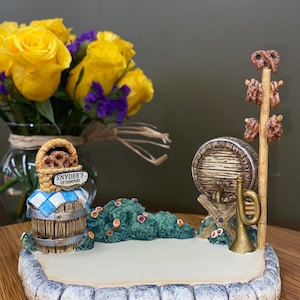 May include: A ceramic miniature scene depicting a German-themed bakery with a barrel, pretzel stand, and a sign that reads "Snyder's of Hanover".