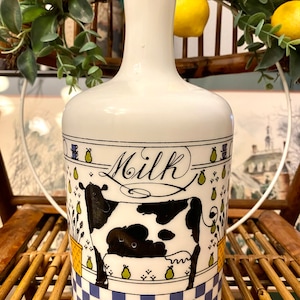 May include: White ceramic milk bottle featuring a black and white cow illustration and the word "Milk" in cursive. The bottle has a blue and white checkered pattern at the base.