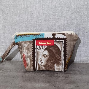 May include: A brown and white fabric cosmetic bag with a zipper closure. The bag features a vintage design with postage stamps and a woman's face. The text "Arte Postale - Post CA" is printed on the bag.