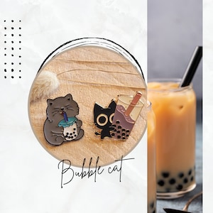 Puede incluir: Dos pines de esmalte dorado con gatos negros bebiendo bubble tea. El texto "Bubble cat" está escrito debajo de los pines. Un vaso de bubble tea con perlas de tapioca está en el fondo.